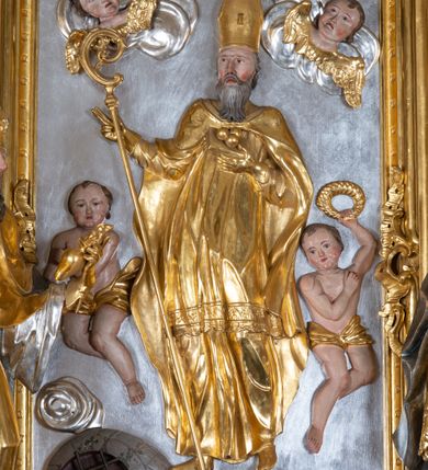 Zdjęcie nr 24: Ołtarz przyścienny, architektoniczny z mensą w formie sarkofagu o wklęsło-wypukłych bokach. W mensie kamienny portatyl. Retabulum jednoosiowe, jednokondygnacyjne ze zwieńczeniem, ustawione na wysokim zdwojonym cokole, zryzalitowanym bo bokach. Nastawa ujęta parą narożnych pilastrów kompozytowych oraz parą kolumn znacznie wysuniętych przed lico ołtarza. Kolumny o kanelowanych trzonach i kompozytowych głowicach podtrzymują faliste belkowanie. Zwieńczenie ujęte po bokach motywami „S” kształtnymi o wolutowo zwiniętych końcach, z których dolne mocno wysunięte do przodu, spoczywają na belkowaniu. Zwieńczenie zamknięte dwoma stykającymi się wolutami. Po bokach retabulum ażurowe uszaki formowane z grzebieni rocaille'owych i motywów „C” kształtnych z rzeźbioną, uskrzydloną główką anielską. 
Pole środkowe w kształcie prostokątnej wnęki z zamknięciem kształtowanym z motywów „C”. We wnęce płaskorzeźba z przedstwieniem św. Mikołaja ujęta podwójną profilowaną ramą. U dołu, po bokach wnęki dwie konsole z ustawionymi pełnoplastycznymi figurami św. Augustyna po lewej oraz św. Marcina (?) po prawej stronie. Ponad kolumnami, po bokach zwieńczenia dwie figury aniołów, powyżej para uskrzydlonych puttów. W zwieńczeniu owalna wnęka z obrazem przedstawiającym św. Sebastiana, ujętym podwójną ramą. 
Ołtarz bogato dekorowany. Mensa zdobiona ornamentem rocaille'owym. Pola cokołów zdobione płycinami kształtowanymi z grzebieni rocaille'owych dekorujących też fryz. Trzony kolumn i pilastrów zdobią podwieszane motywy rocaille'owe przeplatane z gałązkami róż, tworzącymi również rozległy, ażurowy kartusz nad polem środkowym. Boki zwieńczenia ujęte ażurowymi liśćmi akantu. Poniżej wnęki ażurowa, ukośna kratka regencyjna. Boki wnęki i szczyt zwieńczenia dekorowane pełnoplastycznymi rocaille'ami. 
Całość struktury ołtarza pokryta malowaną marmoryzacją w odcieniach zieleni i brązu. Detal architektoniczny złocony, pilastry srebrzone.