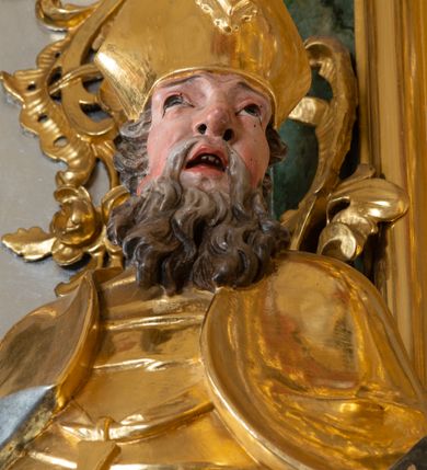 Zdjęcie nr 9: Ołtarz przyścienny, architektoniczny z mensą w formie sarkofagu o wklęsło-wypukłych bokach. W mensie kamienny portatyl. Retabulum jednoosiowe, jednokondygnacyjne ze zwieńczeniem, ustawione na wysokim zdwojonym cokole, zryzalitowanym bo bokach. Nastawa ujęta parą narożnych pilastrów kompozytowych oraz parą kolumn znacznie wysuniętych przed lico ołtarza. Kolumny o kanelowanych trzonach i kompozytowych głowicach podtrzymują faliste belkowanie. Zwieńczenie ujęte po bokach motywami „S” kształtnymi o wolutowo zwiniętych końcach, z których dolne mocno wysunięte do przodu, spoczywają na belkowaniu. Zwieńczenie zamknięte dwoma stykającymi się wolutami. Po bokach retabulum ażurowe uszaki formowane z grzebieni rocaille&#039;owych i motywów „C” kształtnych z rzeźbioną, uskrzydloną główką anielską. 
Pole środkowe w kształcie prostokątnej wnęki z zamknięciem kształtowanym z motywów „C”. We wnęce płaskorzeźba z przedstwieniem św. Mikołaja ujęta podwójną profilowaną ramą. U dołu, po bokach wnęki dwie konsole z ustawionymi pełnoplastycznymi figurami św. Augustyna po lewej oraz św. Marcina (?) po prawej stronie. Ponad kolumnami, po bokach zwieńczenia dwie figury aniołów, powyżej para uskrzydlonych puttów. W zwieńczeniu owalna wnęka z obrazem przedstawiającym św. Sebastiana, ujętym podwójną ramą. 
Ołtarz bogato dekorowany. Mensa zdobiona ornamentem rocaille&#039;owym. Pola cokołów zdobione płycinami kształtowanymi z grzebieni rocaille&#039;owych dekorujących też fryz. Trzony kolumn i pilastrów zdobią podwieszane motywy rocaille&#039;owe przeplatane z gałązkami róż, tworzącymi również rozległy, ażurowy kartusz nad polem środkowym. Boki zwieńczenia ujęte ażurowymi liśćmi akantu. Poniżej wnęki ażurowa, ukośna kratka regencyjna. Boki wnęki i szczyt zwieńczenia dekorowane pełnoplastycznymi rocaille&#039;ami. 
Całość struktury ołtarza pokryta malowaną marmoryzacją w odcieniach zieleni i brązu. Detal architektoniczny złocony, pilastry srebrzone.