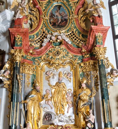 Zdjęcie nr 13: Ołtarz przyścienny, architektoniczny z mensą w formie sarkofagu o wklęsło-wypukłych bokach. W mensie kamienny portatyl. Retabulum jednoosiowe, jednokondygnacyjne ze zwieńczeniem, ustawione na wysokim zdwojonym cokole, zryzalitowanym bo bokach. Nastawa ujęta parą narożnych pilastrów kompozytowych oraz parą kolumn znacznie wysuniętych przed lico ołtarza. Kolumny o kanelowanych trzonach i kompozytowych głowicach podtrzymują faliste belkowanie. Zwieńczenie ujęte po bokach motywami „S” kształtnymi o wolutowo zwiniętych końcach, z których dolne mocno wysunięte do przodu, spoczywają na belkowaniu. Zwieńczenie zamknięte dwoma stykającymi się wolutami. Po bokach retabulum ażurowe uszaki formowane z grzebieni rocaille'owych i motywów „C” kształtnych z rzeźbioną, uskrzydloną główką anielską. 
Pole środkowe w kształcie prostokątnej wnęki z zamknięciem kształtowanym z motywów „C”. We wnęce płaskorzeźba z przedstwieniem św. Mikołaja ujęta podwójną profilowaną ramą. U dołu, po bokach wnęki dwie konsole z ustawionymi pełnoplastycznymi figurami św. Augustyna po lewej oraz św. Marcina (?) po prawej stronie. Ponad kolumnami, po bokach zwieńczenia dwie figury aniołów, powyżej para uskrzydlonych puttów. W zwieńczeniu owalna wnęka z obrazem przedstawiającym św. Sebastiana, ujętym podwójną ramą. 
Ołtarz bogato dekorowany. Mensa zdobiona ornamentem rocaille'owym. Pola cokołów zdobione płycinami kształtowanymi z grzebieni rocaille'owych dekorujących też fryz. Trzony kolumn i pilastrów zdobią podwieszane motywy rocaille'owe przeplatane z gałązkami róż, tworzącymi również rozległy, ażurowy kartusz nad polem środkowym. Boki zwieńczenia ujęte ażurowymi liśćmi akantu. Poniżej wnęki ażurowa, ukośna kratka regencyjna. Boki wnęki i szczyt zwieńczenia dekorowane pełnoplastycznymi rocaille'ami. 
Całość struktury ołtarza pokryta malowaną marmoryzacją w odcieniach zieleni i brązu. Detal architektoniczny złocony, pilastry srebrzone.