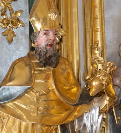 Zdjęcie nr 8: Ołtarz przyścienny, architektoniczny z mensą w formie sarkofagu o wklęsło-wypukłych bokach. W mensie kamienny portatyl. Retabulum jednoosiowe, jednokondygnacyjne ze zwieńczeniem, ustawione na wysokim zdwojonym cokole, zryzalitowanym bo bokach. Nastawa ujęta parą narożnych pilastrów kompozytowych oraz parą kolumn znacznie wysuniętych przed lico ołtarza. Kolumny o kanelowanych trzonach i kompozytowych głowicach podtrzymują faliste belkowanie. Zwieńczenie ujęte po bokach motywami „S” kształtnymi o wolutowo zwiniętych końcach, z których dolne mocno wysunięte do przodu, spoczywają na belkowaniu. Zwieńczenie zamknięte dwoma stykającymi się wolutami. Po bokach retabulum ażurowe uszaki formowane z grzebieni rocaille&#039;owych i motywów „C” kształtnych z rzeźbioną, uskrzydloną główką anielską. 
Pole środkowe w kształcie prostokątnej wnęki z zamknięciem kształtowanym z motywów „C”. We wnęce płaskorzeźba z przedstwieniem św. Mikołaja ujęta podwójną profilowaną ramą. U dołu, po bokach wnęki dwie konsole z ustawionymi pełnoplastycznymi figurami św. Augustyna po lewej oraz św. Marcina (?) po prawej stronie. Ponad kolumnami, po bokach zwieńczenia dwie figury aniołów, powyżej para uskrzydlonych puttów. W zwieńczeniu owalna wnęka z obrazem przedstawiającym św. Sebastiana, ujętym podwójną ramą. 
Ołtarz bogato dekorowany. Mensa zdobiona ornamentem rocaille&#039;owym. Pola cokołów zdobione płycinami kształtowanymi z grzebieni rocaille&#039;owych dekorujących też fryz. Trzony kolumn i pilastrów zdobią podwieszane motywy rocaille&#039;owe przeplatane z gałązkami róż, tworzącymi również rozległy, ażurowy kartusz nad polem środkowym. Boki zwieńczenia ujęte ażurowymi liśćmi akantu. Poniżej wnęki ażurowa, ukośna kratka regencyjna. Boki wnęki i szczyt zwieńczenia dekorowane pełnoplastycznymi rocaille&#039;ami. 
Całość struktury ołtarza pokryta malowaną marmoryzacją w odcieniach zieleni i brązu. Detal architektoniczny złocony, pilastry srebrzone.