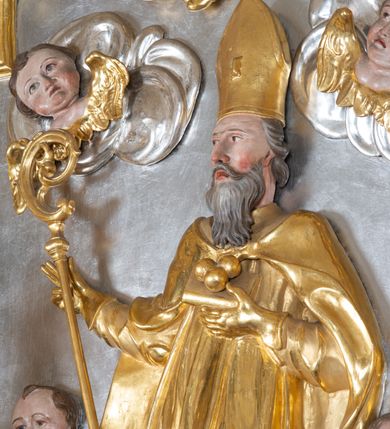 Zdjęcie nr 28: Ołtarz przyścienny, architektoniczny z mensą w formie sarkofagu o wklęsło-wypukłych bokach. W mensie kamienny portatyl. Retabulum jednoosiowe, jednokondygnacyjne ze zwieńczeniem, ustawione na wysokim zdwojonym cokole, zryzalitowanym bo bokach. Nastawa ujęta parą narożnych pilastrów kompozytowych oraz parą kolumn znacznie wysuniętych przed lico ołtarza. Kolumny o kanelowanych trzonach i kompozytowych głowicach podtrzymują faliste belkowanie. Zwieńczenie ujęte po bokach motywami „S” kształtnymi o wolutowo zwiniętych końcach, z których dolne mocno wysunięte do przodu, spoczywają na belkowaniu. Zwieńczenie zamknięte dwoma stykającymi się wolutami. Po bokach retabulum ażurowe uszaki formowane z grzebieni rocaille'owych i motywów „C” kształtnych z rzeźbioną, uskrzydloną główką anielską. 
Pole środkowe w kształcie prostokątnej wnęki z zamknięciem kształtowanym z motywów „C”. We wnęce płaskorzeźba z przedstwieniem św. Mikołaja ujęta podwójną profilowaną ramą. U dołu, po bokach wnęki dwie konsole z ustawionymi pełnoplastycznymi figurami św. Augustyna po lewej oraz św. Marcina (?) po prawej stronie. Ponad kolumnami, po bokach zwieńczenia dwie figury aniołów, powyżej para uskrzydlonych puttów. W zwieńczeniu owalna wnęka z obrazem przedstawiającym św. Sebastiana, ujętym podwójną ramą. 
Ołtarz bogato dekorowany. Mensa zdobiona ornamentem rocaille'owym. Pola cokołów zdobione płycinami kształtowanymi z grzebieni rocaille'owych dekorujących też fryz. Trzony kolumn i pilastrów zdobią podwieszane motywy rocaille'owe przeplatane z gałązkami róż, tworzącymi również rozległy, ażurowy kartusz nad polem środkowym. Boki zwieńczenia ujęte ażurowymi liśćmi akantu. Poniżej wnęki ażurowa, ukośna kratka regencyjna. Boki wnęki i szczyt zwieńczenia dekorowane pełnoplastycznymi rocaille'ami. 
Całość struktury ołtarza pokryta malowaną marmoryzacją w odcieniach zieleni i brązu. Detal architektoniczny złocony, pilastry srebrzone.