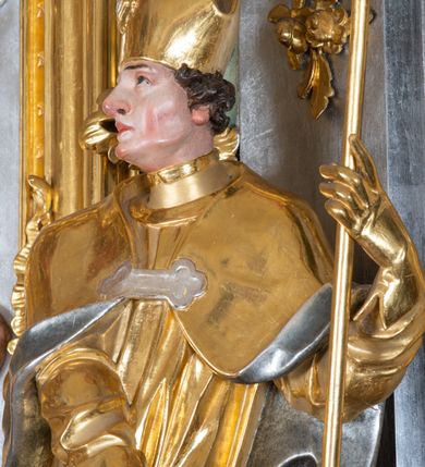 Zdjęcie nr 20: Ołtarz przyścienny, architektoniczny z mensą w formie sarkofagu o wklęsło-wypukłych bokach. W mensie kamienny portatyl. Retabulum jednoosiowe, jednokondygnacyjne ze zwieńczeniem, ustawione na wysokim zdwojonym cokole, zryzalitowanym bo bokach. Nastawa ujęta parą narożnych pilastrów kompozytowych oraz parą kolumn znacznie wysuniętych przed lico ołtarza. Kolumny o kanelowanych trzonach i kompozytowych głowicach podtrzymują faliste belkowanie. Zwieńczenie ujęte po bokach motywami „S” kształtnymi o wolutowo zwiniętych końcach, z których dolne mocno wysunięte do przodu, spoczywają na belkowaniu. Zwieńczenie zamknięte dwoma stykającymi się wolutami. Po bokach retabulum ażurowe uszaki formowane z grzebieni rocaille'owych i motywów „C” kształtnych z rzeźbioną, uskrzydloną główką anielską. 
Pole środkowe w kształcie prostokątnej wnęki z zamknięciem kształtowanym z motywów „C”. We wnęce płaskorzeźba z przedstwieniem św. Mikołaja ujęta podwójną profilowaną ramą. U dołu, po bokach wnęki dwie konsole z ustawionymi pełnoplastycznymi figurami św. Augustyna po lewej oraz św. Marcina (?) po prawej stronie. Ponad kolumnami, po bokach zwieńczenia dwie figury aniołów, powyżej para uskrzydlonych puttów. W zwieńczeniu owalna wnęka z obrazem przedstawiającym św. Sebastiana, ujętym podwójną ramą. 
Ołtarz bogato dekorowany. Mensa zdobiona ornamentem rocaille'owym. Pola cokołów zdobione płycinami kształtowanymi z grzebieni rocaille'owych dekorujących też fryz. Trzony kolumn i pilastrów zdobią podwieszane motywy rocaille'owe przeplatane z gałązkami róż, tworzącymi również rozległy, ażurowy kartusz nad polem środkowym. Boki zwieńczenia ujęte ażurowymi liśćmi akantu. Poniżej wnęki ażurowa, ukośna kratka regencyjna. Boki wnęki i szczyt zwieńczenia dekorowane pełnoplastycznymi rocaille'ami. 
Całość struktury ołtarza pokryta malowaną marmoryzacją w odcieniach zieleni i brązu. Detal architektoniczny złocony, pilastry srebrzone.