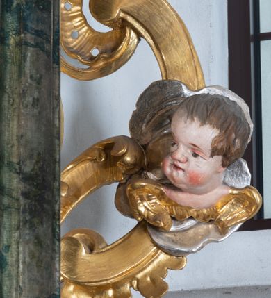 Zdjęcie nr 27: Ołtarz przyścienny, architektoniczny z mensą w formie sarkofagu o wklęsło-wypukłych bokach. W mensie kamienny portatyl. Retabulum jednoosiowe, jednokondygnacyjne ze zwieńczeniem, ustawione na wysokim zdwojonym cokole, zryzalitowanym bo bokach. Nastawa ujęta parą narożnych pilastrów kompozytowych oraz parą kolumn znacznie wysuniętych przed lico ołtarza. Kolumny o kanelowanych trzonach i kompozytowych głowicach podtrzymują faliste belkowanie. Zwieńczenie ujęte po bokach motywami „S” kształtnymi o wolutowo zwiniętych końcach, z których dolne mocno wysunięte do przodu, spoczywają na belkowaniu. Zwieńczenie zamknięte dwoma stykającymi się wolutami. Po bokach retabulum ażurowe uszaki formowane z grzebieni rocaille'owych i motywów „C” kształtnych z rzeźbioną, uskrzydloną główką anielską. 
Pole środkowe w kształcie prostokątnej wnęki z zamknięciem kształtowanym z motywów „C”. We wnęce płaskorzeźba z przedstwieniem św. Mikołaja ujęta podwójną profilowaną ramą. U dołu, po bokach wnęki dwie konsole z ustawionymi pełnoplastycznymi figurami św. Augustyna po lewej oraz św. Marcina (?) po prawej stronie. Ponad kolumnami, po bokach zwieńczenia dwie figury aniołów, powyżej para uskrzydlonych puttów. W zwieńczeniu owalna wnęka z obrazem przedstawiającym św. Sebastiana, ujętym podwójną ramą. 
Ołtarz bogato dekorowany. Mensa zdobiona ornamentem rocaille'owym. Pola cokołów zdobione płycinami kształtowanymi z grzebieni rocaille'owych dekorujących też fryz. Trzony kolumn i pilastrów zdobią podwieszane motywy rocaille'owe przeplatane z gałązkami róż, tworzącymi również rozległy, ażurowy kartusz nad polem środkowym. Boki zwieńczenia ujęte ażurowymi liśćmi akantu. Poniżej wnęki ażurowa, ukośna kratka regencyjna. Boki wnęki i szczyt zwieńczenia dekorowane pełnoplastycznymi rocaille'ami. 
Całość struktury ołtarza pokryta malowaną marmoryzacją w odcieniach zieleni i brązu. Detal architektoniczny złocony, pilastry srebrzone.