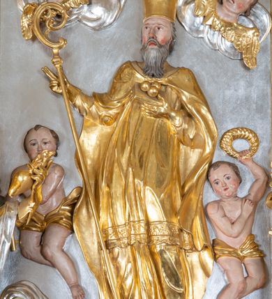 Zdjęcie nr 29: Ołtarz przyścienny, architektoniczny z mensą w formie sarkofagu o wklęsło-wypukłych bokach. W mensie kamienny portatyl. Retabulum jednoosiowe, jednokondygnacyjne ze zwieńczeniem, ustawione na wysokim zdwojonym cokole, zryzalitowanym bo bokach. Nastawa ujęta parą narożnych pilastrów kompozytowych oraz parą kolumn znacznie wysuniętych przed lico ołtarza. Kolumny o kanelowanych trzonach i kompozytowych głowicach podtrzymują faliste belkowanie. Zwieńczenie ujęte po bokach motywami „S” kształtnymi o wolutowo zwiniętych końcach, z których dolne mocno wysunięte do przodu, spoczywają na belkowaniu. Zwieńczenie zamknięte dwoma stykającymi się wolutami. Po bokach retabulum ażurowe uszaki formowane z grzebieni rocaille&#039;owych i motywów „C” kształtnych z rzeźbioną, uskrzydloną główką anielską. 
Pole środkowe w kształcie prostokątnej wnęki z zamknięciem kształtowanym z motywów „C”. We wnęce płaskorzeźba z przedstwieniem św. Mikołaja ujęta podwójną profilowaną ramą. U dołu, po bokach wnęki dwie konsole z ustawionymi pełnoplastycznymi figurami św. Augustyna po lewej oraz św. Marcina (?) po prawej stronie. Ponad kolumnami, po bokach zwieńczenia dwie figury aniołów, powyżej para uskrzydlonych puttów. W zwieńczeniu owalna wnęka z obrazem przedstawiającym św. Sebastiana, ujętym podwójną ramą. 
Ołtarz bogato dekorowany. Mensa zdobiona ornamentem rocaille&#039;owym. Pola cokołów zdobione płycinami kształtowanymi z grzebieni rocaille&#039;owych dekorujących też fryz. Trzony kolumn i pilastrów zdobią podwieszane motywy rocaille&#039;owe przeplatane z gałązkami róż, tworzącymi również rozległy, ażurowy kartusz nad polem środkowym. Boki zwieńczenia ujęte ażurowymi liśćmi akantu. Poniżej wnęki ażurowa, ukośna kratka regencyjna. Boki wnęki i szczyt zwieńczenia dekorowane pełnoplastycznymi rocaille&#039;ami. 
Całość struktury ołtarza pokryta malowaną marmoryzacją w odcieniach zieleni i brązu. Detal architektoniczny złocony, pilastry srebrzone.