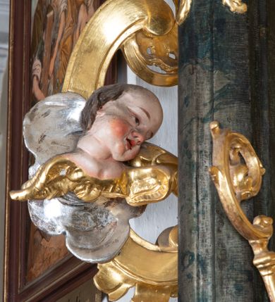 Zdjęcie nr 26: Ołtarz przyścienny, architektoniczny z mensą w formie sarkofagu o wklęsło-wypukłych bokach. W mensie kamienny portatyl. Retabulum jednoosiowe, jednokondygnacyjne ze zwieńczeniem, ustawione na wysokim zdwojonym cokole, zryzalitowanym bo bokach. Nastawa ujęta parą narożnych pilastrów kompozytowych oraz parą kolumn znacznie wysuniętych przed lico ołtarza. Kolumny o kanelowanych trzonach i kompozytowych głowicach podtrzymują faliste belkowanie. Zwieńczenie ujęte po bokach motywami „S” kształtnymi o wolutowo zwiniętych końcach, z których dolne mocno wysunięte do przodu, spoczywają na belkowaniu. Zwieńczenie zamknięte dwoma stykającymi się wolutami. Po bokach retabulum ażurowe uszaki formowane z grzebieni rocaille'owych i motywów „C” kształtnych z rzeźbioną, uskrzydloną główką anielską. 
Pole środkowe w kształcie prostokątnej wnęki z zamknięciem kształtowanym z motywów „C”. We wnęce płaskorzeźba z przedstwieniem św. Mikołaja ujęta podwójną profilowaną ramą. U dołu, po bokach wnęki dwie konsole z ustawionymi pełnoplastycznymi figurami św. Augustyna po lewej oraz św. Marcina (?) po prawej stronie. Ponad kolumnami, po bokach zwieńczenia dwie figury aniołów, powyżej para uskrzydlonych puttów. W zwieńczeniu owalna wnęka z obrazem przedstawiającym św. Sebastiana, ujętym podwójną ramą. 
Ołtarz bogato dekorowany. Mensa zdobiona ornamentem rocaille'owym. Pola cokołów zdobione płycinami kształtowanymi z grzebieni rocaille'owych dekorujących też fryz. Trzony kolumn i pilastrów zdobią podwieszane motywy rocaille'owe przeplatane z gałązkami róż, tworzącymi również rozległy, ażurowy kartusz nad polem środkowym. Boki zwieńczenia ujęte ażurowymi liśćmi akantu. Poniżej wnęki ażurowa, ukośna kratka regencyjna. Boki wnęki i szczyt zwieńczenia dekorowane pełnoplastycznymi rocaille'ami. 
Całość struktury ołtarza pokryta malowaną marmoryzacją w odcieniach zieleni i brązu. Detal architektoniczny złocony, pilastry srebrzone.