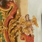 Zdjęcie nr 1: Na wolutowo-wywiniętych przyczółkach zwieńczenia usadzona jest para aniołów. Anioł po lewej stronie ukazany jest w pozycji siedzącej, zwrócony trzy czwarte w lewo z lewą nogą podwiniętą, a prawą opuszczoną w dół; lewą rękę składa na piersi, a prawą wyciąga do przodu; głowę ma uniesioną do góry. Twarz szeroka z wyraźnie zaznaczonym podbródkiem, szerokim na końcu nosem i dużym oczami. Włosy krótkie i kędzierzawe, ciemnobrązowe z loczkiem zawiniętym nad czołem. Anioł ubrany jest z złoconą tunikę ze srebrzoną podszewką, odsłaniającą nagi tors i prawą nogę powyżej uda. U ramion ma duże, złocone i rozpięte na boki skrzydła. Po prawej stronie ołtarza znajduje się drugi anioł o antytetycznym układzie sylwetki z rękami ugiętymi w łokciach i rozłożonymi na boki oraz głową skierowaną delikatnie w lewo. Szaty, skrzydła i twarz analogiczne, jak u pierwszego anioła. 

Wyżej, na wolutowym zakończeniu boków zwieńczenia usadzona jest para małych aniołków z niewielkimi skrzydłami, opasanymi w biodrach złotą przepaską. Aniołki ukazane są w ruchu o esowato wygiętej sylwetce, z jedną nogą uniesioną do góry i rękami odwiedzionymi na jeden bok. Twarze szerokie z pulchnymi policzkami, nosami o szerokich końcach i dużymi oczami, włosy krótkie i kędzierzawe. Polichromia w odsłoniętych partiach ciała naturalistyczna, szaty i skrzydła złocone. 
