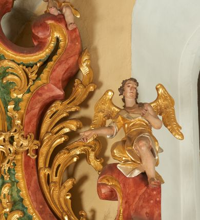 Zdjęcie nr 1: Na wolutowo-wywiniętych przyczółkach zwieńczenia usadzona jest para aniołów. Anioł po lewej stronie ukazany jest w pozycji siedzącej, zwrócony trzy czwarte w lewo z lewą nogą podwiniętą, a prawą opuszczoną w dół; lewą rękę składa na piersi, a prawą wyciąga do przodu; głowę ma uniesioną do góry. Twarz szeroka z wyraźnie zaznaczonym podbródkiem, szerokim na końcu nosem i dużym oczami. Włosy krótkie i kędzierzawe, ciemnobrązowe z loczkiem zawiniętym nad czołem. Anioł ubrany jest z złoconą tunikę ze srebrzoną podszewką, odsłaniającą nagi tors i prawą nogę powyżej uda. U ramion ma duże, złocone i rozpięte na boki skrzydła. Po prawej stronie ołtarza znajduje się drugi anioł o antytetycznym układzie sylwetki z rękami ugiętymi w łokciach i rozłożonymi na boki oraz głową skierowaną delikatnie w lewo. Szaty, skrzydła i twarz analogiczne, jak u pierwszego anioła. 

Wyżej, na wolutowym zakończeniu boków zwieńczenia usadzona jest para małych aniołków z niewielkimi skrzydłami, opasanymi w biodrach złotą przepaską. Aniołki ukazane są w ruchu o esowato wygiętej sylwetce, z jedną nogą uniesioną do góry i rękami odwiedzionymi na jeden bok. Twarze szerokie z pulchnymi policzkami, nosami o szerokich końcach i dużymi oczami, włosy krótkie i kędzierzawe. Polichromia w odsłoniętych partiach ciała naturalistyczna, szaty i skrzydła złocone. 
