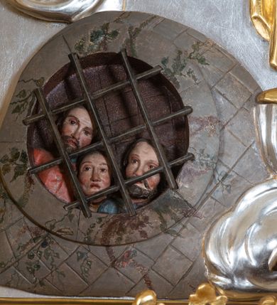 Zdjęcie nr 4: Płaskorzeźba w kształcie prostokąta zamkniętego wklęsło-wypukłym łukiem zdobionym grzebieniami rocaille’owymi pośrodku. W polu, na jednolicie srebrnym tle ukazany św. Mikołaj, stojący na wolutowo zwiniętych obłokach. Święty zwrócony delikatnie w prawą stronę, ręce uniesione na wysokości piersi i ugięte w łokciach, w lewej przylegającej do piersi trzyma zamkniętą księgę z trzema złotymi kulami, a w prawej pastorał. Twarz świętego pociągła, okolona długą i siwą brodą, oczy niewielkie, nos długi o prostym profilu, usta rozchylone o opadających kącikach. Święty ubrany jest w szaty biskupie: albę, rokietę oraz kapę spiętą pod szyją. Na głowie ma infułę, na dłoniach rękawiczki, na stopach pełne buty. Po bokach świętego ukazane są dwa aniołki ze złotymi szarfami na biodrach. Anioł po prawej stronie trzyma w uniesionej lewej ręce wieniec, anioł po lewej stronie w opuszczonej prawej ręce trzyma klucz. W lewym dolnym rogu ukazano więzienie, w którym za kratą są trzej mężczyźni. W górnej części uskrzydlone główki aniołków na obłokach. Polichromia w odsłoniętych partiach ciała naturalistyczna, szaty i atrybuty złocone, detale srebrzone.