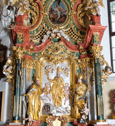 Zdjęcie nr 7: Płaskorzeźba w kształcie prostokąta zamkniętego wklęsło-wypukłym łukiem zdobionym grzebieniami rocaille’owymi pośrodku. W polu, na jednolicie srebrnym tle ukazany św. Mikołaj, stojący na wolutowo zwiniętych obłokach. Święty zwrócony delikatnie w prawą stronę, ręce uniesione na wysokości piersi i ugięte w łokciach, w lewej przylegającej do piersi trzyma zamkniętą księgę z trzema złotymi kulami, a w prawej pastorał. Twarz świętego pociągła, okolona długą i siwą brodą, oczy niewielkie, nos długi o prostym profilu, usta rozchylone o opadających kącikach. Święty ubrany jest w szaty biskupie: albę, rokietę oraz kapę spiętą pod szyją. Na głowie ma infułę, na dłoniach rękawiczki, na stopach pełne buty. Po bokach świętego ukazane są dwa aniołki ze złotymi szarfami na biodrach. Anioł po prawej stronie trzyma w uniesionej lewej ręce wieniec, anioł po lewej stronie w opuszczonej prawej ręce trzyma klucz. W lewym dolnym rogu ukazano więzienie, w którym za kratą są trzej mężczyźni. W górnej części uskrzydlone główki aniołków na obłokach. Polichromia w odsłoniętych partiach ciała naturalistyczna, szaty i atrybuty złocone, detale srebrzone.