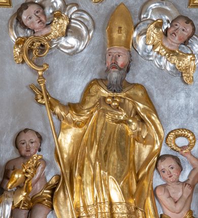 Zdjęcie nr 1: Płaskorzeźba w kształcie prostokąta zamkniętego wklęsło-wypukłym łukiem zdobionym grzebieniami rocaille’owymi pośrodku. W polu, na jednolicie srebrnym tle ukazany św. Mikołaj, stojący na wolutowo zwiniętych obłokach. Święty zwrócony delikatnie w prawą stronę, ręce uniesione na wysokości piersi i ugięte w łokciach, w lewej przylegającej do piersi trzyma zamkniętą księgę z trzema złotymi kulami, a w prawej pastorał. Twarz świętego pociągła, okolona długą i siwą brodą, oczy niewielkie, nos długi o prostym profilu, usta rozchylone o opadających kącikach. Święty ubrany jest w szaty biskupie: albę, rokietę oraz kapę spiętą pod szyją. Na głowie ma infułę, na dłoniach rękawiczki, na stopach pełne buty. Po bokach świętego ukazane są dwa aniołki ze złotymi szarfami na biodrach. Anioł po prawej stronie trzyma w uniesionej lewej ręce wieniec, anioł po lewej stronie w opuszczonej prawej ręce trzyma klucz. W lewym dolnym rogu ukazano więzienie, w którym za kratą są trzej mężczyźni. W górnej części uskrzydlone główki aniołków na obłokach. Polichromia w odsłoniętych partiach ciała naturalistyczna, szaty i atrybuty złocone, detale srebrzone.