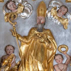 Zdjęcie nr 1: Płaskorzeźba w kształcie prostokąta zamkniętego wklęsło-wypukłym łukiem zdobionym grzebieniami rocaille’owymi pośrodku. W polu, na jednolicie srebrnym tle ukazany św. Mikołaj, stojący na wolutowo zwiniętych obłokach. Święty zwrócony delikatnie w prawą stronę, ręce uniesione na wysokości piersi i ugięte w łokciach, w lewej przylegającej do piersi trzyma zamkniętą księgę z trzema złotymi kulami, a w prawej pastorał. Twarz świętego pociągła, okolona długą i siwą brodą, oczy niewielkie, nos długi o prostym profilu, usta rozchylone o opadających kącikach. Święty ubrany jest w szaty biskupie: albę, rokietę oraz kapę spiętą pod szyją. Na głowie ma infułę, na dłoniach rękawiczki, na stopach pełne buty. Po bokach świętego ukazane są dwa aniołki ze złotymi szarfami na biodrach. Anioł po prawej stronie trzyma w uniesionej lewej ręce wieniec, anioł po lewej stronie w opuszczonej prawej ręce trzyma klucz. W lewym dolnym rogu ukazano więzienie, w którym za kratą są trzej mężczyźni. W górnej części uskrzydlone główki aniołków na obłokach. Polichromia w odsłoniętych partiach ciała naturalistyczna, szaty i atrybuty złocone, detale srebrzone.