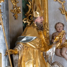 Zdjęcie nr 1: Rzeźba św. Augustyna ścięta z tyłu, drążona, ustawiona na prostopadłościennym cokole. Święty ukazany frontalnie w silnym kontrapoście z lewą nogą ugiętą w kolanie. W prawej dłoni trzyma pastorał, a w lewej ugiętej w łokciu gorejące serce. Twarz szeroka o wyraźnie zaznaczonych kościach policzkowych, dużych oczach szeroko rozstawionych na boki i charakterystycznym dużym nosie, policzki silnie zarumienione, usta szeroko otwarte. Włosy krótkie, brązowe, zawinięte w loki do tyłu, broda bujna i długa. Święty ubrany jest długą złoconą albę ze srebrzonymi rękawami, mucet i złotą kapę o srebrzonej podszewce. Na głowie ma założoną złoconą infułę, nosi również złocone rękawice i pełne buty, a na szyi ma zawieszony krzyż pektoralny. U nóg świętego, po prawej stronie siedzi mały chłopczyk z rękami rozłożonymi szeroko na boki, w prawej trzyma łyżkę. Twarz pełna z dużym, charakterystycznym dla twórcy nosem, silnie zarumienonymi policzkami, okolona krótkimi ciemnobrązowymi włosami. Ubrany jest jedynie w złoconą chustę, opasającą biodra. Polichromia naturalistyczna w odsłoniętych partiach ciała; szaty i atrybuty złocone oraz srebrzone.

