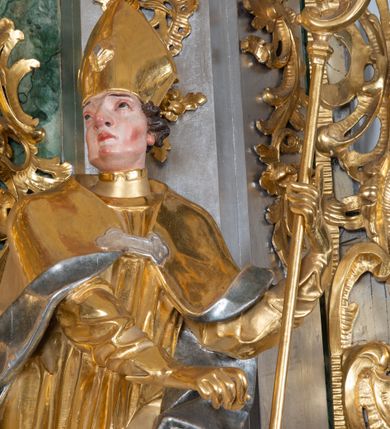 Zdjęcie nr 6: Rzeźba św. Marcina ścięta z tyłu, drążona, ustawiona na prostopadłościennym cokole. Święty ukazany jest frontalnie w silnym kontrapoście. Ręce ma ugięte w łokciach i skierowane  na lewy bok, trzyma w nich pastorał. Twarz szeroka o wyraźnie zaznaczonych kościach policzkowych, dużych oczach szeroko rozstawionych na boki i charakterystycznym dużym nosie, policzki silnie zarumienione. Włosy krótkie, ciemnobrązowe, zawinięte w loki do tyłu. Święty ubrany jest długą złoconą albę oraz rokietę i złotą kapę o srebrzonej podszewce, spiętą na piersi szeroką klamrą. Na głowie ma założoną złoconą infułę, nosi również złocone rękawice i pełne buty.  U nóg świętego, po lewej stronie klęczy na prawym kolanie żebrak wyciągający kapelusz na jałmużnę. Sylwetka drobna. Twarz szeroka o wyraźnie podkreślonych kościach policzkowych z osadzonymi szeroko oczami, otwartymi ustami i charakterystycznym dużym nosem. Głowa okolona krótką i siwą brodą oraz włosami z łysiną nad czołem. Żebrak ubrany jest w srebrzoną szatę, odsłaniającą lewe, nagie ramię. Polichromia naturalistyczna w odsłoniętych partiach ciała; szaty i atrybuty złocone oraz srebrzone.


