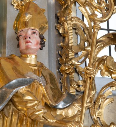 Zdjęcie nr 5: Rzeźba św. Marcina ścięta z tyłu, drążona, ustawiona na prostopadłościennym cokole. Święty ukazany jest frontalnie w silnym kontrapoście. Ręce ma ugięte w łokciach i skierowane  na lewy bok, trzyma w nich pastorał. Twarz szeroka o wyraźnie zaznaczonych kościach policzkowych, dużych oczach szeroko rozstawionych na boki i charakterystycznym dużym nosie, policzki silnie zarumienione. Włosy krótkie, ciemnobrązowe, zawinięte w loki do tyłu. Święty ubrany jest długą złoconą albę oraz rokietę i złotą kapę o srebrzonej podszewce, spiętą na piersi szeroką klamrą. Na głowie ma założoną złoconą infułę, nosi również złocone rękawice i pełne buty.  U nóg świętego, po lewej stronie klęczy na prawym kolanie żebrak wyciągający kapelusz na jałmużnę. Sylwetka drobna. Twarz szeroka o wyraźnie podkreślonych kościach policzkowych z osadzonymi szeroko oczami, otwartymi ustami i charakterystycznym dużym nosem. Głowa okolona krótką i siwą brodą oraz włosami z łysiną nad czołem. Żebrak ubrany jest w srebrzoną szatę, odsłaniającą lewe, nagie ramię. Polichromia naturalistyczna w odsłoniętych partiach ciała; szaty i atrybuty złocone oraz srebrzone.


