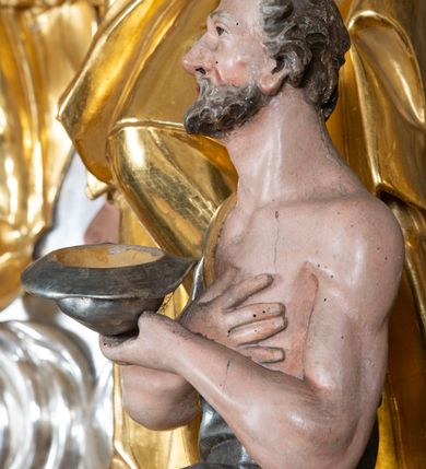 Zdjęcie nr 8: Rzeźba św. Marcina ścięta z tyłu, drążona, ustawiona na prostopadłościennym cokole. Święty ukazany jest frontalnie w silnym kontrapoście. Ręce ma ugięte w łokciach i skierowane  na lewy bok, trzyma w nich pastorał. Twarz szeroka o wyraźnie zaznaczonych kościach policzkowych, dużych oczach szeroko rozstawionych na boki i charakterystycznym dużym nosie, policzki silnie zarumienione. Włosy krótkie, ciemnobrązowe, zawinięte w loki do tyłu. Święty ubrany jest długą złoconą albę oraz rokietę i złotą kapę o srebrzonej podszewce, spiętą na piersi szeroką klamrą. Na głowie ma założoną złoconą infułę, nosi również złocone rękawice i pełne buty.  U nóg świętego, po lewej stronie klęczy na prawym kolanie żebrak wyciągający kapelusz na jałmużnę. Sylwetka drobna. Twarz szeroka o wyraźnie podkreślonych kościach policzkowych z osadzonymi szeroko oczami, otwartymi ustami i charakterystycznym dużym nosem. Głowa okolona krótką i siwą brodą oraz włosami z łysiną nad czołem. Żebrak ubrany jest w srebrzoną szatę, odsłaniającą lewe, nagie ramię. Polichromia naturalistyczna w odsłoniętych partiach ciała; szaty i atrybuty złocone oraz srebrzone.


