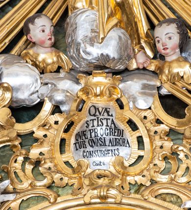 Zdjęcie nr 34: Nastawa ołtarzowa architektoniczna, trójosiowa z bramkami, jednokondygnacyjna ze zwieńczeniem. Ołtarz wysunięty do przodu, drewniany, sarkofagowy, wklęsło-wypukły, zwężający się ku dołowi, na profilowanym cokole, ujęty spływami wolutowymi; dekorowany w narożach palmetami, na froncie rocaille&#039;owy kartusz z malowanymi skrzyżowanymi kluczami w polu. Mensa drewniana z silnie wysuniętą, profilowaną krawędzią, z portatylem z piaskowca. 

Tabernakulum w formie prostopadłościennej skrzynki o wypukłej ściance frontowej, ujęte z przodu ukośnie dostawionymi pilastrami na cokołach z trzonami w formie wolut, dźwigającymi niepełne belkowanie, opasujące je z trzech stron, nad ścianką frontową przyjmujące falistą formę zwieńczoną rocaille&#039;m. Drzwiczki przyjmują kształt stojącego prostokąta zamkniętego łukiem półkolistym nadwieszonym, ujęte profilowaną ramą; w polu płaskorzeźbione przedstawienie kielicha z hostią w glorii na fakturowanym tle. Na belkowaniu, na osi podpór ustawiona jest para rocaille&#039;owych lichtarzy. Tron wystawienia architektoniczny, trójosiowy; oś środkowa ponad tabernakulum w postaci ażurowej niszy z cokołem na pasyjkę, tylna ścianka utworzona z rocaille&#039;u z kłosami zbóż; flankowana pilastrami zwieńczonymi falistym gzymsem, dźwigającymi ażurową, zamkniętą koronę. Podpory przechodzą w spływy wolutowe, ujmujące tabernakulum, na których końcach siedzi para adorujących puttów. Osie boczne są ujęte po bokach pilastrami dźwigającymi imposty podtrzymywane przez rocaille w partii trzonów oraz płonące wazony; pola wypełnione ażurowym rocaille&#039;m i palmetami przechodzącymi w podwójne lichtarze.

Retabulum na planie półowalu, osie boczne ustawione prostopadle do środkowej. Oś środkowa posadowiona na wysokim, dwustrefowym cokole. W pierwszej strefie cokołu prostokątna płycina z napisem: „Z INICJATYWY / KS. PROBOSZCZA ROMANA GORCZYŃSKIEGO / W LATACH 2001 - 2006 / WYKONANO REMONT KOŚCIOŁA I KONSERWACJĘ / WYPOSAŻENIA”. W drugiej strefie cokołu, pośrodku płycina w formie leżącego prostokąta o fantazyjnie wyciętych bokach z malowanym kartuszem herbowym, dzielonym na cztery pola. W lewym górnym polu kartusza umieszczony jest orzeł, poniżej dwójka dzieci w łódce, a po prawej stronie tarczy, w prawym górnym polu dwójka dzieci w łódce, poniżej orzeł. U spodu napis z chronostychem: „EREXIT, AC EXORNAVIT HONORIBVS SANCTI PETRI, / AC PAVLI APOSTOLI ILLVSTRISSIMVS LIBER BAR I WNBS(?) IOANELLY / A TOLVANA PROPPII X I”. Klejnotem jest hełm z pióropuszem zakończony półpostacią rycerza w zbroi, zaś labry mają formę bujnej wici akantowej.  Ryzality cokołów ozdobione są z trzech stron rocaille&#039;owymi płycinami; skrajne konsole rocaille&#039;m i różanymi gałązkami. Oś główna flankowana parą kolumn ustawioną na tle zwielokrotnionych pilastrów. Podpory dźwigają pełne belkowanie, przerwane nad polem środkowym, wyłamane na ich osi oraz przerwany wolutowy przyczółek. W polu środkowym podwójna, profilowana rama w formie stojącego prostokąta zamkniętego łukiem nadwieszonym z uskokami z obrazem św. Piotra i św. Pawła; flankowana przez figury św. Szymona po lewej stronie i św. Judy Tadeusza po prawej, ustawionymi na rocaille&#039;owych wspornikach. Ramy obrazu, trzony wewnętrznych pilastrów i pola za środkową parą figur dekorowane rocaille&#039;m. 

Zwieńczenie w formie zbliżonej do stojącego prostokąta, ujęte spływami wolutowymi, zamknięte falistym gzymsem przechodzącym w antytetycznie zwrócone woluty. Całość zwieńczona gołębicą Ducha Świętego w glorii promienistej. W polu rzeźbiarska grupa Koronacji Marii w glorii promienistej, pośród obłoków i uskrzydlonych główek anielskich. Dwa flankujące grupę putta  na obłokach trzymają banderole z napisem: „FILIA DEI PATRIS” i „MATER DEI FILII”, a kolejne dwa usadzone na szczycie: „VENI CORONABERIS” oraz „VENI SPONSA MEA”. Poniżej zwieńczenia w ażurowym kartuszu znajduje się napis: „QVA E/ST ISTA / QVAE PROGREDI/TUR QVASI AURORA / CONSURGENS / Cant 6.”. Na przyczółku siedzące figury św. Joachima po lewej stronie i św. Józefa po prawej, na gzymsie flankującym pole środkowe dwa putta trzymające słońce i księżyc, na zewnętrznych osiach dwa adorujące anioły. Spływy zwieńczenia dekorowane strzępiastymi liśćmi palmowymi, muszlą i rocaille’m. 

Osie boczne ustawione są na wysokim cokole, od zewnątrz ujęte pilastrami przechodzącymi w woluty, od wewnątrz sąsiadują z osią środkową. Pośrodku bramki z drzwiczkami zakończonymi gzymsem przyjmującym formę cokołu dla figur. Drzwiczki w formie stojącego prostokątna zamkniętego łukiem segmentowym nadwieszonym; osie na skraju ujęte konsolami stanowiącymi cokoły dla podpór. Ryzalit bramek zdobiony płyciną ze strzępiastym grzebieniem; drzwiczki z płyciną z rocaille&#039;m i kwiatowym zwisem na wstążce; powyższy cokół ujęty liśćmi palmowymi i rocaille&#039;m. Kondygnacja flankowana na skraju kolumną ustawioną na tle pilastru, które podtrzymują wyłamany gzyms zakończony wolutami ujmującymi szczyt o falistym przebiegu, na którym ustawiony jest duży płomienisty wazon. W polach ustawione są figury: po lewej się Jakuba Starszego, a po prawej św. Filipa. Na zwieńczeniach usadzone dwie figury aniołów. 

Struktura w całości marmoryzowana w kolorze zielono-brązowym; ornamentyka, kolumny złocone, detale srebrzone. Wszystkie kolumny w jednej trzeciej wysokości przedzielone rocaille’m, a fryz z elementami dekoracji rokokowej. Tabernakulum i tron wystawienia w całości złocone.

