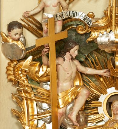 Zdjęcie nr 4: W polu zwieńczenia znajduje się rzeźbiarska grupa przedstawiająca Koronację Matki Boskiej na tle promienistej glorii, pośród obłoków i aniołków. W centrum kompozycji ukazana jest Matka Boska w pozycji klęczącej ze skrzyżowanymi na piersi rękami. Ma szeroką twarz z wyraźnie zaznaczonym podbródkiem, delikatnie rozchylonymi ustami, szerokim na końcu nosem i migdałowymi oczami. Jej głowę okala srebrno-złoty, okrągły nimb, od którego odchodzą wiązki promieni o nierównej długości. Ubrana jest w złocone szaty: długą suknię, płaszcz i welon na głowie. Ma ciemnobrązowe włosy, schowane pod welonem. Po jej bokach na srebrnych obłokach zasiadają: po prawej Bóg Ojciec, a po lewej Jezus. Obaj zwróceni są trzy czwarte ku środkowi, w jednej ręce trzymają swoje atrybuty: Bóg Ojciec berło, a Chrystus wysoki krzyż; a drugą wyciągniętą w bok podtrzymują nad głową Marii pełną koronę. Bóg Ojciec ubrany jest w złoconą suknię oraz złocony płaszcz. Ma podłużną twarz z lekko rozchylonymi ustami, siwe włosy oraz długą i siwą brodę, skręconą w dwa długie pukle. Jezus ma lewą nogę ugiętą w kolanie i uniesioną wyżej. Twarz pociągła, okolona krótkim zarostem; włosy długie, kręcone i przylegające do głowy. Przez biodra ma przepasane krótkie i złocone perizonium. Ciało szczupłe, o wyraźnej linii mostka i żeber, z zaznaczonymi śladami męki. W dolnej części przedstawienia grupę flankują dwa putta na obłokach, które trzymają banderole z napisami: „FILIA DEI PATRIS” i „MATER DEI FILII”, a kolejne dwa putta usadzone na szczycie: „VENI CORONABERIS” oraz „VENI SPONSA MEA”. Poniżej zwieńczenia w ażurowym kartuszu znajduje się napis: „QVA E/ST ISTA / QVAE PROGREDI/TUR QVASI AURORA / CONSURGENS / Cant 6.”. Na gzymsie flankującym pole środkowe dwa putta trzymają słońce i księżyc. Całość zdobiona jest ponadto sześcioma główkami anielskimi. Na szczycie zwieńczenia znajduje się gołębica Ducha Świętego na tle promienistej glorii. Draperia szat traktowana jest miękko. Spływy zwieńczenia dekorowane są strzępiastymi liśćmi palmowymi, muszlą i rocaille’m.Polichromia naturalistyczna w odsłoniętych partiach ciała; szaty i atrybuty złocone, obłoki srebrzone.