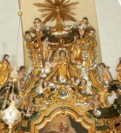 Zdjęcie nr 2: W polu zwieńczenia znajduje się rzeźbiarska grupa przedstawiająca Koronację Matki Boskiej na tle promienistej glorii, pośród obłoków i aniołków. W centrum kompozycji ukazana jest Matka Boska w pozycji klęczącej ze skrzyżowanymi na piersi rękami. Ma szeroką twarz z wyraźnie zaznaczonym podbródkiem, delikatnie rozchylonymi ustami, szerokim na końcu nosem i migdałowymi oczami. Jej głowę okala srebrno-złoty, okrągły nimb, od którego odchodzą wiązki promieni o nierównej długości. Ubrana jest w złocone szaty: długą suknię, płaszcz i welon na głowie. Ma ciemnobrązowe włosy, schowane pod welonem. Po jej bokach na srebrnych obłokach zasiadają: po prawej Bóg Ojciec, a po lewej Jezus. Obaj zwróceni są trzy czwarte ku środkowi, w jednej ręce trzymają swoje atrybuty: Bóg Ojciec berło, a Chrystus wysoki krzyż; a drugą wyciągniętą w bok podtrzymują nad głową Marii pełną koronę. Bóg Ojciec ubrany jest w złoconą suknię oraz złocony płaszcz. Ma podłużną twarz z lekko rozchylonymi ustami, siwe włosy oraz długą i siwą brodę, skręconą w dwa długie pukle. Jezus ma lewą nogę ugiętą w kolanie i uniesioną wyżej. Twarz pociągła, okolona krótkim zarostem; włosy długie, kręcone i przylegające do głowy. Przez biodra ma przepasane krótkie i złocone perizonium. Ciało szczupłe, o wyraźnej linii mostka i żeber, z zaznaczonymi śladami męki. W dolnej części przedstawienia grupę flankują dwa putta na obłokach, które trzymają banderole z napisami: „FILIA DEI PATRIS” i „MATER DEI FILII”, a kolejne dwa putta usadzone na szczycie: „VENI CORONABERIS” oraz „VENI SPONSA MEA”. Poniżej zwieńczenia w ażurowym kartuszu znajduje się napis: „QVA E/ST ISTA / QVAE PROGREDI/TUR QVASI AURORA / CONSURGENS / Cant 6.”. Na gzymsie flankującym pole środkowe dwa putta trzymają słońce i księżyc. Całość zdobiona jest ponadto sześcioma główkami anielskimi. Na szczycie zwieńczenia znajduje się gołębica Ducha Świętego na tle promienistej glorii. Draperia szat traktowana jest miękko. Spływy zwieńczenia dekorowane są strzępiastymi liśćmi palmowymi, muszlą i rocaille’m.Polichromia naturalistyczna w odsłoniętych partiach ciała; szaty i atrybuty złocone, obłoki srebrzone.