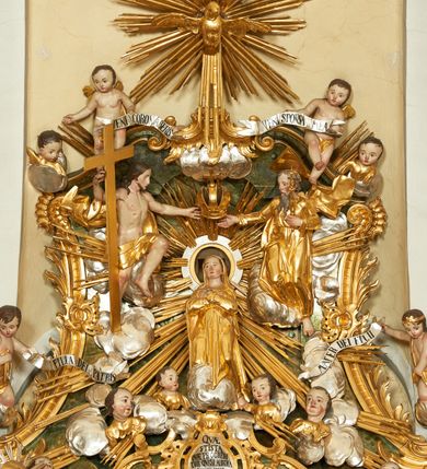 Zdjęcie nr 1: W polu zwieńczenia znajduje się rzeźbiarska grupa przedstawiająca Koronację Matki Boskiej na tle promienistej glorii, pośród obłoków i aniołków. W centrum kompozycji ukazana jest Matka Boska w pozycji klęczącej ze skrzyżowanymi na piersi rękami. Ma szeroką twarz z wyraźnie zaznaczonym podbródkiem, delikatnie rozchylonymi ustami, szerokim na końcu nosem i migdałowymi oczami. Jej głowę okala srebrno-złoty, okrągły nimb, od którego odchodzą wiązki promieni o nierównej długości. Ubrana jest w złocone szaty: długą suknię, płaszcz i welon na głowie. Ma ciemnobrązowe włosy, schowane pod welonem. Po jej bokach na srebrnych obłokach zasiadają: po prawej Bóg Ojciec, a po lewej Jezus. Obaj zwróceni są trzy czwarte ku środkowi, w jednej ręce trzymają swoje atrybuty: Bóg Ojciec berło, a Chrystus wysoki krzyż; a drugą wyciągniętą w bok podtrzymują nad głową Marii pełną koronę. Bóg Ojciec ubrany jest w złoconą suknię oraz złocony płaszcz. Ma podłużną twarz z lekko rozchylonymi ustami, siwe włosy oraz długą i siwą brodę, skręconą w dwa długie pukle. Jezus ma lewą nogę ugiętą w kolanie i uniesioną wyżej. Twarz pociągła, okolona krótkim zarostem; włosy długie, kręcone i przylegające do głowy. Przez biodra ma przepasane krótkie i złocone perizonium. Ciało szczupłe, o wyraźnej linii mostka i żeber, z zaznaczonymi śladami męki. W dolnej części przedstawienia grupę flankują dwa putta na obłokach, które trzymają banderole z napisami: „FILIA DEI PATRIS” i „MATER DEI FILII”, a kolejne dwa putta usadzone na szczycie: „VENI CORONABERIS” oraz „VENI SPONSA MEA”. Poniżej zwieńczenia w ażurowym kartuszu znajduje się napis: „QVA E/ST ISTA / QVAE PROGREDI/TUR QVASI AURORA / CONSURGENS / Cant 6.”. Na gzymsie flankującym pole środkowe dwa putta trzymają słońce i księżyc. Całość zdobiona jest ponadto sześcioma główkami anielskimi. Na szczycie zwieńczenia znajduje się gołębica Ducha Świętego na tle promienistej glorii. Draperia szat traktowana jest miękko. Spływy zwieńczenia dekorowane są strzępiastymi liśćmi palmowymi, muszlą i rocaille’m.Polichromia naturalistyczna w odsłoniętych partiach ciała; szaty i atrybuty złocone, obłoki srebrzone.