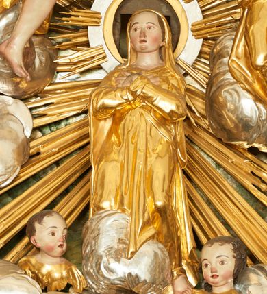 Zdjęcie nr 3: W polu zwieńczenia znajduje się rzeźbiarska grupa przedstawiająca Koronację Matki Boskiej na tle promienistej glorii, pośród obłoków i aniołków. W centrum kompozycji ukazana jest Matka Boska w pozycji klęczącej ze skrzyżowanymi na piersi rękami. Ma szeroką twarz z wyraźnie zaznaczonym podbródkiem, delikatnie rozchylonymi ustami, szerokim na końcu nosem i migdałowymi oczami. Jej głowę okala srebrno-złoty, okrągły nimb, od którego odchodzą wiązki promieni o nierównej długości. Ubrana jest w złocone szaty: długą suknię, płaszcz i welon na głowie. Ma ciemnobrązowe włosy, schowane pod welonem. Po jej bokach na srebrnych obłokach zasiadają: po prawej Bóg Ojciec, a po lewej Jezus. Obaj zwróceni są trzy czwarte ku środkowi, w jednej ręce trzymają swoje atrybuty: Bóg Ojciec berło, a Chrystus wysoki krzyż; a drugą wyciągniętą w bok podtrzymują nad głową Marii pełną koronę. Bóg Ojciec ubrany jest w złoconą suknię oraz złocony płaszcz. Ma podłużną twarz z lekko rozchylonymi ustami, siwe włosy oraz długą i siwą brodę, skręconą w dwa długie pukle. Jezus ma lewą nogę ugiętą w kolanie i uniesioną wyżej. Twarz pociągła, okolona krótkim zarostem; włosy długie, kręcone i przylegające do głowy. Przez biodra ma przepasane krótkie i złocone perizonium. Ciało szczupłe, o wyraźnej linii mostka i żeber, z zaznaczonymi śladami męki. W dolnej części przedstawienia grupę flankują dwa putta na obłokach, które trzymają banderole z napisami: „FILIA DEI PATRIS” i „MATER DEI FILII”, a kolejne dwa putta usadzone na szczycie: „VENI CORONABERIS” oraz „VENI SPONSA MEA”. Poniżej zwieńczenia w ażurowym kartuszu znajduje się napis: „QVA E/ST ISTA / QVAE PROGREDI/TUR QVASI AURORA / CONSURGENS / Cant 6.”. Na gzymsie flankującym pole środkowe dwa putta trzymają słońce i księżyc. Całość zdobiona jest ponadto sześcioma główkami anielskimi. Na szczycie zwieńczenia znajduje się gołębica Ducha Świętego na tle promienistej glorii. Draperia szat traktowana jest miękko. Spływy zwieńczenia dekorowane są strzępiastymi liśćmi palmowymi, muszlą i rocaille’m.Polichromia naturalistyczna w odsłoniętych partiach ciała; szaty i atrybuty złocone, obłoki srebrzone.