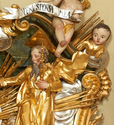 Zdjęcie nr 6: W polu zwieńczenia znajduje się rzeźbiarska grupa przedstawiająca Koronację Matki Boskiej na tle promienistej glorii, pośród obłoków i aniołków. W centrum kompozycji ukazana jest Matka Boska w pozycji klęczącej ze skrzyżowanymi na piersi rękami. Ma szeroką twarz z wyraźnie zaznaczonym podbródkiem, delikatnie rozchylonymi ustami, szerokim na końcu nosem i migdałowymi oczami. Jej głowę okala srebrno-złoty, okrągły nimb, od którego odchodzą wiązki promieni o nierównej długości. Ubrana jest w złocone szaty: długą suknię, płaszcz i welon na głowie. Ma ciemnobrązowe włosy, schowane pod welonem. Po jej bokach na srebrnych obłokach zasiadają: po prawej Bóg Ojciec, a po lewej Jezus. Obaj zwróceni są trzy czwarte ku środkowi, w jednej ręce trzymają swoje atrybuty: Bóg Ojciec berło, a Chrystus wysoki krzyż; a drugą wyciągniętą w bok podtrzymują nad głową Marii pełną koronę. Bóg Ojciec ubrany jest w złoconą suknię oraz złocony płaszcz. Ma podłużną twarz z lekko rozchylonymi ustami, siwe włosy oraz długą i siwą brodę, skręconą w dwa długie pukle. Jezus ma lewą nogę ugiętą w kolanie i uniesioną wyżej. Twarz pociągła, okolona krótkim zarostem; włosy długie, kręcone i przylegające do głowy. Przez biodra ma przepasane krótkie i złocone perizonium. Ciało szczupłe, o wyraźnej linii mostka i żeber, z zaznaczonymi śladami męki. W dolnej części przedstawienia grupę flankują dwa putta na obłokach, które trzymają banderole z napisami: „FILIA DEI PATRIS” i „MATER DEI FILII”, a kolejne dwa putta usadzone na szczycie: „VENI CORONABERIS” oraz „VENI SPONSA MEA”. Poniżej zwieńczenia w ażurowym kartuszu znajduje się napis: „QVA E/ST ISTA / QVAE PROGREDI/TUR QVASI AURORA / CONSURGENS / Cant 6.”. Na gzymsie flankującym pole środkowe dwa putta trzymają słońce i księżyc. Całość zdobiona jest ponadto sześcioma główkami anielskimi. Na szczycie zwieńczenia znajduje się gołębica Ducha Świętego na tle promienistej glorii. Draperia szat traktowana jest miękko. Spływy zwieńczenia dekorowane są strzępiastymi liśćmi palmowymi, muszlą i rocaille’m.Polichromia naturalistyczna w odsłoniętych partiach ciała; szaty i atrybuty złocone, obłoki srebrzone.