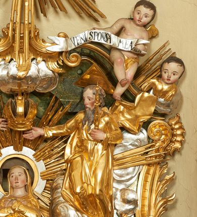 Zdjęcie nr 5: W polu zwieńczenia znajduje się rzeźbiarska grupa przedstawiająca Koronację Matki Boskiej na tle promienistej glorii, pośród obłoków i aniołków. W centrum kompozycji ukazana jest Matka Boska w pozycji klęczącej ze skrzyżowanymi na piersi rękami. Ma szeroką twarz z wyraźnie zaznaczonym podbródkiem, delikatnie rozchylonymi ustami, szerokim na końcu nosem i migdałowymi oczami. Jej głowę okala srebrno-złoty, okrągły nimb, od którego odchodzą wiązki promieni o nierównej długości. Ubrana jest w złocone szaty: długą suknię, płaszcz i welon na głowie. Ma ciemnobrązowe włosy, schowane pod welonem. Po jej bokach na srebrnych obłokach zasiadają: po prawej Bóg Ojciec, a po lewej Jezus. Obaj zwróceni są trzy czwarte ku środkowi, w jednej ręce trzymają swoje atrybuty: Bóg Ojciec berło, a Chrystus wysoki krzyż; a drugą wyciągniętą w bok podtrzymują nad głową Marii pełną koronę. Bóg Ojciec ubrany jest w złoconą suknię oraz złocony płaszcz. Ma podłużną twarz z lekko rozchylonymi ustami, siwe włosy oraz długą i siwą brodę, skręconą w dwa długie pukle. Jezus ma lewą nogę ugiętą w kolanie i uniesioną wyżej. Twarz pociągła, okolona krótkim zarostem; włosy długie, kręcone i przylegające do głowy. Przez biodra ma przepasane krótkie i złocone perizonium. Ciało szczupłe, o wyraźnej linii mostka i żeber, z zaznaczonymi śladami męki. W dolnej części przedstawienia grupę flankują dwa putta na obłokach, które trzymają banderole z napisami: „FILIA DEI PATRIS” i „MATER DEI FILII”, a kolejne dwa putta usadzone na szczycie: „VENI CORONABERIS” oraz „VENI SPONSA MEA”. Poniżej zwieńczenia w ażurowym kartuszu znajduje się napis: „QVA E/ST ISTA / QVAE PROGREDI/TUR QVASI AURORA / CONSURGENS / Cant 6.”. Na gzymsie flankującym pole środkowe dwa putta trzymają słońce i księżyc. Całość zdobiona jest ponadto sześcioma główkami anielskimi. Na szczycie zwieńczenia znajduje się gołębica Ducha Świętego na tle promienistej glorii. Draperia szat traktowana jest miękko. Spływy zwieńczenia dekorowane są strzępiastymi liśćmi palmowymi, muszlą i rocaille’m.Polichromia naturalistyczna w odsłoniętych partiach ciała; szaty i atrybuty złocone, obłoki srebrzone.