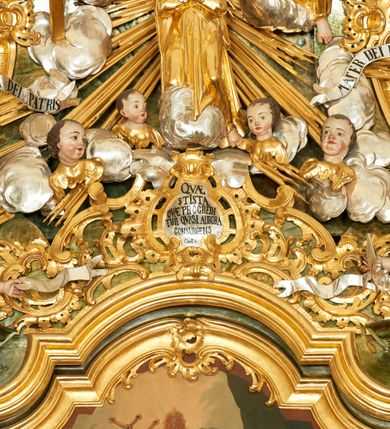Zdjęcie nr 7: W polu zwieńczenia znajduje się rzeźbiarska grupa przedstawiająca Koronację Matki Boskiej na tle promienistej glorii, pośród obłoków i aniołków. W centrum kompozycji ukazana jest Matka Boska w pozycji klęczącej ze skrzyżowanymi na piersi rękami. Ma szeroką twarz z wyraźnie zaznaczonym podbródkiem, delikatnie rozchylonymi ustami, szerokim na końcu nosem i migdałowymi oczami. Jej głowę okala srebrno-złoty, okrągły nimb, od którego odchodzą wiązki promieni o nierównej długości. Ubrana jest w złocone szaty: długą suknię, płaszcz i welon na głowie. Ma ciemnobrązowe włosy, schowane pod welonem. Po jej bokach na srebrnych obłokach zasiadają: po prawej Bóg Ojciec, a po lewej Jezus. Obaj zwróceni są trzy czwarte ku środkowi, w jednej ręce trzymają swoje atrybuty: Bóg Ojciec berło, a Chrystus wysoki krzyż; a drugą wyciągniętą w bok podtrzymują nad głową Marii pełną koronę. Bóg Ojciec ubrany jest w złoconą suknię oraz złocony płaszcz. Ma podłużną twarz z lekko rozchylonymi ustami, siwe włosy oraz długą i siwą brodę, skręconą w dwa długie pukle. Jezus ma lewą nogę ugiętą w kolanie i uniesioną wyżej. Twarz pociągła, okolona krótkim zarostem; włosy długie, kręcone i przylegające do głowy. Przez biodra ma przepasane krótkie i złocone perizonium. Ciało szczupłe, o wyraźnej linii mostka i żeber, z zaznaczonymi śladami męki. W dolnej części przedstawienia grupę flankują dwa putta na obłokach, które trzymają banderole z napisami: „FILIA DEI PATRIS” i „MATER DEI FILII”, a kolejne dwa putta usadzone na szczycie: „VENI CORONABERIS” oraz „VENI SPONSA MEA”. Poniżej zwieńczenia w ażurowym kartuszu znajduje się napis: „QVA E/ST ISTA / QVAE PROGREDI/TUR QVASI AURORA / CONSURGENS / Cant 6.”. Na gzymsie flankującym pole środkowe dwa putta trzymają słońce i księżyc. Całość zdobiona jest ponadto sześcioma główkami anielskimi. Na szczycie zwieńczenia znajduje się gołębica Ducha Świętego na tle promienistej glorii. Draperia szat traktowana jest miękko. Spływy zwieńczenia dekorowane są strzępiastymi liśćmi palmowymi, muszlą i rocaille’m.Polichromia naturalistyczna w odsłoniętych partiach ciała; szaty i atrybuty złocone, obłoki srebrzone.