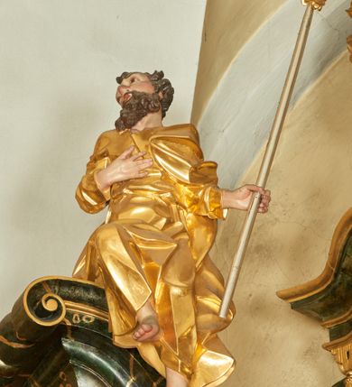 Zdjęcie nr 1: Rzeźba św. Józefa ścięta z tyłu, drążona, ustawiona na przyczółku ołtarza. Święty zwrócony trzy czwarte w prawo, ukazany w pozycji siedzącej z prawą nogą ugiętą w kolanie i delikatnie uniesioną do góry, w lewej dłoni trzyma kwitnącą laskę, a prawą ma złożoną na piersi. Twarz szeroka o wyraźnie zaznaczonych kościach policzkowych, dużych oczach i nosie, policzki i nos silnie zarumienione, twarz okolona krótką brodą. Włosy długie, ciemnobrązowe spływające na plecy i lewe ramię. Święty ubrany jest w długą suknię z długimi i wąskimi rękawami, drapowaną w grube i ostro łamane fałdy z dekoltem w kształcie litery „V”. Polichromia naturalistyczna w odsłoniętych partiach ciała; szaty złocone, laska o srebrzonym drzewcu.

