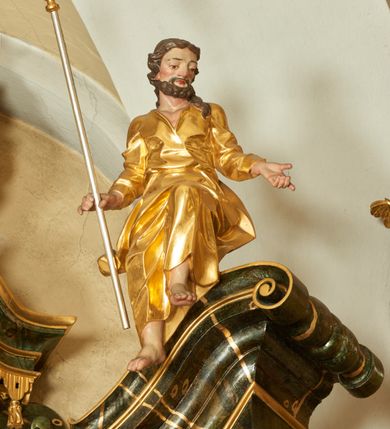 Zdjęcie nr 1: Rzeźba św. Joachima ścięta z tyłu, drążona, ustawiona na przyczółku ołtarza. Święty zwrócony trzy czwarte w prawo, ukazany w pozycji siedzącej z lewą nogą ugiętą w kolanie i delikatnie uniesioną do góry, ręce szeroko rozłożone na boki, w prawej trzyma kij pasterski o zakończeniu w kształcie szufli. Twarz szeroka o wyraźnie zaznaczonych kościach policzkowych, dużych oczach i nosie, policzki i nos silnie zarumienione, twarz okolona krótką brodą. Włosy długie, ciemnobrązowe spływające na plecy i lewe ramię. Święty ubrany jest w długą suknię z długimi i wąskimi rękawami, zawiniętymi na mankietach, drapowaną w grube i ostro łamane fałdy z dekoltem w kształcie litery „V”. Polichromia naturalistyczna w odsłoniętych partiach ciała; szaty złocone, szufla złocona o srebrzonym drzewcu.
