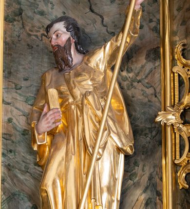 Zdjęcie nr 2: Rzeźba św. Filipa ścięta z tyłu, drążona, ustawiona na prostopadłościennej kostce. Święty ukazany frontalnie w silnym kontrapoście z prawą nogą ugiętą w kolanie. W lewej, uniesionej do góry ręce trzyma krzyż, a w prawej przytrzymuje do boku księgę. Twarz szeroka o wyraźnie zaznaczonych kościach policzkowych, dużych oczach i delikatnie rozchylonych ustach, policzki silnie zarumienione, twarz okolona długą brodą. Włosy długie, ciemnobrązowe spływające na plecy i czoło. Święty ubrany jest w suknię sięgającą do połowy łydek z długimi i wąskimi rękawami, zawiniętymi na mankietach, drapowaną w grube i miękkie fałdy. Na prawe ramię ma narzucony długi płaszcz, przewiązany diagonalnie na torsie wstążką. Polichromia naturalistyczna w odsłoniętych partiach ciała; szaty i atrybuty złocone.
