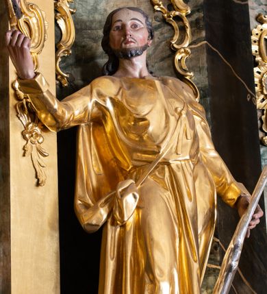 Zdjęcie nr 1: Rzeźba św. Judy Tadeusza ścięta z tyłu, drążona, ustawiona na kostce. Święty ukazany frontalnie w silnym kontrapoście z prawą nogą ugiętą w kolanie. W prawej, uniesionej do góry ręce trzyma medalion z wizerunkiem Chrystusa, a w lewej opuszczonej wzdłuż ciała podtrzymuje pałkę. Twarz szeroka o wyraźnie zaznaczonych kościach policzkowych, dużych oczach i delikatnie rozchylonych ustach, policzki silnie zarumienione, twarz okolona krótką brodą. Włosy długie, ciemnobrązowe spływające na plecy. Święty ubrany jest w suknię sięgającą za kolana z długimi i wąskimi rękawami, zawiniętymi na mankietach, drapowaną w grube i miękkie fałdy. Na lewe ramię narzucony długi płaszcz, przewiązany diagonalnie na torsie wstążką. Polichromia naturalistyczna w odsłoniętych partiach ciała; szaty i atrybuty złocone, pałka srebrzona.
