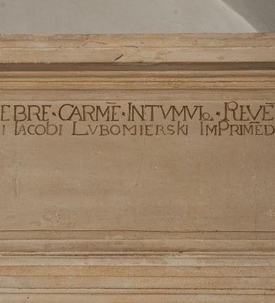 Zdjęcie nr 17: Nagrobek utworzony z dwóch położonych obok siebie płyt w formie leżącego prostokąta, w profilowanych, uszatych obramieniach z płaskorzeźbionymi, leżącymi postaciami Stanisława Lubomirskiego (po lewej) i Jakuba Lubomirskiego (po prawej). Postać po lewej leży na prawym boku, z ręką ugiętą i podpierającą głowę, zaś lewą ułożoną wzdłuż ciała, z buzdyganem w dłoni przyozdobionej pierścieniem, z ugiętą lewą nogą. Jego twarz jest pociągła, nos wydatny, ale krótki, oczy zamknięte. Włosy ma krótkie, lekko kręcone. Ubrany jest w zbroję typu husarskiego, składającą się z napierśnika z dwoma folgami brzusznymi pod szyją zakończonego wysokim kołnierzem, z naramienników, nałokietników, z sześciofolgowych taszek, nakolanników, nagolenników i butów. Na piersi zawieszony ma łańcuch. Pod nim leży hełm z pióropuszem i długi miecz. W prawym, górnym rogu kartusz z owalnym wypukłym polem z godłem herbu Drużyna i syglami w polu &quot;S&quot; i &quot;L&quot;. Nad nim kamienna tablica. 
Na prawej płycie ukazano Jakuba Lubomirskiego, leżącego na lewym boku, na ugiętej, lewej ręce i prawej ułożonej wzdłuż ciała, z księgą pod dłonią. Jego twarz jest pociągła, nos krótki, oczy zamknięte. Twarz okala długa, falista broda i wąsy, na głowie włosy sięgające uszu. Postać jest ubrana w albę i komżę. Nad płytą kamienna tablica z inskrypcją: „FVNEBRE CARME(NI) INTVMVLO REVE(REN)/DI D(OMI)NI IACOBI LVBOMIERSKI IMPRIME(N)DV(M). 
Nagrobek wieńczy czteropolowy kartusz z herbami: Drużyna, Odrowąż (Ogończyk), Warnia (Rak) i Gryf. Nad kartuszem hełm z koroną z zawieszonym u szyi krzyżem kawalerskim i ujęty labrami. 
