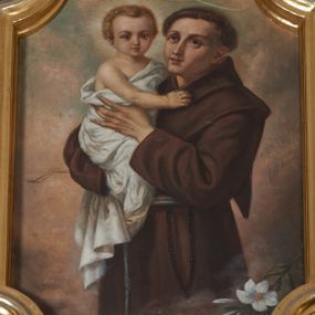 Zdjęcie nr 1: Obraz w kształcie stojącego prostokąta zamkniętego łukiem nadwieszonym z uskokiem, ujęty złoconą, profilowaną ramą. W centrum kompozycji przedstawienie ukazanego w półpostaci, zwróconego w trzech czwartych w prawo św. Antoniego trzymającego na ręku Dzieciątko Jezus. Święty ukazany jako mężczyzna o twarzy pozbawionej zarostu, z tonsurą na głowie, ubrany w brązowy habit przepasany sznurem. U jego boku widoczny różaniec. Święty Antoni oburącz podtrzymuje jasnowłosego Jezusa. Dzieciątko owinięte białą tkaniną. Ponad głową świętego aureola, wokół głowy Dzieciątka nimb promienisty. W dolnej części kompozycji widoczny kwiat białej lilii. Tło jednolicie żółto-brązowe. Kolorystyka obrazu ciemna, ciepła z dominacją brązu.