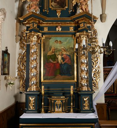 Zdjęcie nr 2: Ołtarz przyścienny, architektoniczny, dwukondygnacyjny, jednoosiowy. Mensa prostopadłościenna, antepedium haftowane z motywem serca gorejącego ujętego winnymi gronami oraz kłosami. Na  mensie architektoniczne, ujęte kolumnami tabernakulum z płaskorzeźbionym kielichem i hostią na drzwiczkach. W pierwszej kondygnacji obraz w kształcie stojącego prostokąta o uskokowo ściętych, górnych narożach z przedstawieniem św. Anny Samotrzeć (na zasuwie obraz „Najświętsze Serce Jezusa”) ujęty umieszczonymi na postumentach, kręconymi, złoconymi  i srebrzonymi kolumnami o kompozytowych kapitelach ustawionymi na tle kanelowanych pilastrów. Podpory dźwigają pełne, wyłamane belkowanie zwieńczone wydatnym, profilowanym gzymsem. Na osiach podpór pełnoplastyczne figury aniołów na obłokach. Fryz dekorowany rozetami oraz uskrzydloną główką anielską. W drugiej kondygnacji obraz św. Franciszka w ramie zamkniętej łukiem półkolistym wyniesionym, z uskokiem. Szczyt w formie przerwanego przyczółka, na osi którego krzyż. Całość nastawy ujęta ażurowymi uszami uformowanymi  z suchego akantu oraz wstęgi. Dekoracja akantowa, rozety, pola wypełnione kratą. Struktura nastawy w odcieniu ciemnej, morskiej zieleni, ornament złocony i srebrzony. 