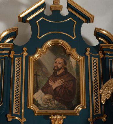 Zdjęcie nr 14: Ołtarz przyścienny, architektoniczny, dwukondygnacyjny, jednoosiowy. Mensa prostopadłościenna, antepedium haftowane z motywem serca gorejącego ujętego winnymi gronami oraz kłosami. Na  mensie architektoniczne, ujęte kolumnami tabernakulum z płaskorzeźbionym kielichem i hostią na drzwiczkach. W pierwszej kondygnacji obraz w kształcie stojącego prostokąta o uskokowo ściętych, górnych narożach z przedstawieniem św. Anny Samotrzeć (na zasuwie obraz „Najświętsze Serce Jezusa”) ujęty umieszczonymi na postumentach, kręconymi, złoconymi  i srebrzonymi kolumnami o kompozytowych kapitelach ustawionymi na tle kanelowanych pilastrów. Podpory dźwigają pełne, wyłamane belkowanie zwieńczone wydatnym, profilowanym gzymsem. Na osiach podpór pełnoplastyczne figury aniołów na obłokach. Fryz dekorowany rozetami oraz uskrzydloną główką anielską. W drugiej kondygnacji obraz św. Franciszka w ramie zamkniętej łukiem półkolistym wyniesionym, z uskokiem. Szczyt w formie przerwanego przyczółka, na osi którego krzyż. Całość nastawy ujęta ażurowymi uszami uformowanymi  z suchego akantu oraz wstęgi. Dekoracja akantowa, rozety, pola wypełnione kratą. Struktura nastawy w odcieniu ciemnej, morskiej zieleni, ornament złocony i srebrzony. 