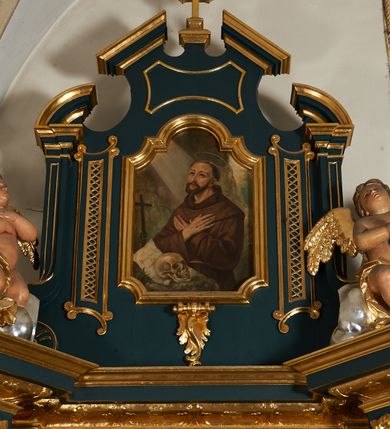Zdjęcie nr 12: Ołtarz przyścienny, architektoniczny, dwukondygnacyjny, jednoosiowy. Mensa prostopadłościenna, antepedium haftowane z motywem serca gorejącego ujętego winnymi gronami oraz kłosami. Na  mensie architektoniczne, ujęte kolumnami tabernakulum z płaskorzeźbionym kielichem i hostią na drzwiczkach. W pierwszej kondygnacji obraz w kształcie stojącego prostokąta o uskokowo ściętych, górnych narożach z przedstawieniem św. Anny Samotrzeć (na zasuwie obraz „Najświętsze Serce Jezusa”) ujęty umieszczonymi na postumentach, kręconymi, złoconymi  i srebrzonymi kolumnami o kompozytowych kapitelach ustawionymi na tle kanelowanych pilastrów. Podpory dźwigają pełne, wyłamane belkowanie zwieńczone wydatnym, profilowanym gzymsem. Na osiach podpór pełnoplastyczne figury aniołów na obłokach. Fryz dekorowany rozetami oraz uskrzydloną główką anielską. W drugiej kondygnacji obraz św. Franciszka w ramie zamkniętej łukiem półkolistym wyniesionym, z uskokiem. Szczyt w formie przerwanego przyczółka, na osi którego krzyż. Całość nastawy ujęta ażurowymi uszami uformowanymi  z suchego akantu oraz wstęgi. Dekoracja akantowa, rozety, pola wypełnione kratą. Struktura nastawy w odcieniu ciemnej, morskiej zieleni, ornament złocony i srebrzony. 