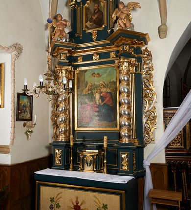 Zdjęcie nr 27: Ołtarz przyścienny, architektoniczny, dwukondygnacyjny, jednoosiowy. Mensa prostopadłościenna, antepedium haftowane z motywem serca gorejącego ujętego winnymi gronami oraz kłosami. Na  mensie architektoniczne, ujęte kolumnami tabernakulum z płaskorzeźbionym kielichem i hostią na drzwiczkach. W pierwszej kondygnacji obraz w kształcie stojącego prostokąta o uskokowo ściętych, górnych narożach z przedstawieniem św. Anny Samotrzeć (na zasuwie obraz „Najświętsze Serce Jezusa”) ujęty umieszczonymi na postumentach, kręconymi, złoconymi  i srebrzonymi kolumnami o kompozytowych kapitelach ustawionymi na tle kanelowanych pilastrów. Podpory dźwigają pełne, wyłamane belkowanie zwieńczone wydatnym, profilowanym gzymsem. Na osiach podpór pełnoplastyczne figury aniołów na obłokach. Fryz dekorowany rozetami oraz uskrzydloną główką anielską. W drugiej kondygnacji obraz św. Franciszka w ramie zamkniętej łukiem półkolistym wyniesionym, z uskokiem. Szczyt w formie przerwanego przyczółka, na osi którego krzyż. Całość nastawy ujęta ażurowymi uszami uformowanymi  z suchego akantu oraz wstęgi. Dekoracja akantowa, rozety, pola wypełnione kratą. Struktura nastawy w odcieniu ciemnej, morskiej zieleni, ornament złocony i srebrzony. 