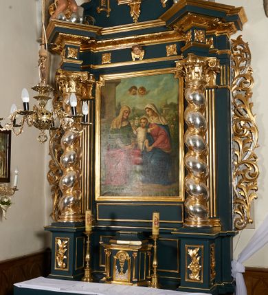 Zdjęcie nr 29: Ołtarz przyścienny, architektoniczny, dwukondygnacyjny, jednoosiowy. Mensa prostopadłościenna, antepedium haftowane z motywem serca gorejącego ujętego winnymi gronami oraz kłosami. Na  mensie architektoniczne, ujęte kolumnami tabernakulum z płaskorzeźbionym kielichem i hostią na drzwiczkach. W pierwszej kondygnacji obraz w kształcie stojącego prostokąta o uskokowo ściętych, górnych narożach z przedstawieniem św. Anny Samotrzeć (na zasuwie obraz „Najświętsze Serce Jezusa”) ujęty umieszczonymi na postumentach, kręconymi, złoconymi  i srebrzonymi kolumnami o kompozytowych kapitelach ustawionymi na tle kanelowanych pilastrów. Podpory dźwigają pełne, wyłamane belkowanie zwieńczone wydatnym, profilowanym gzymsem. Na osiach podpór pełnoplastyczne figury aniołów na obłokach. Fryz dekorowany rozetami oraz uskrzydloną główką anielską. W drugiej kondygnacji obraz św. Franciszka w ramie zamkniętej łukiem półkolistym wyniesionym, z uskokiem. Szczyt w formie przerwanego przyczółka, na osi którego krzyż. Całość nastawy ujęta ażurowymi uszami uformowanymi  z suchego akantu oraz wstęgi. Dekoracja akantowa, rozety, pola wypełnione kratą. Struktura nastawy w odcieniu ciemnej, morskiej zieleni, ornament złocony i srebrzony. 