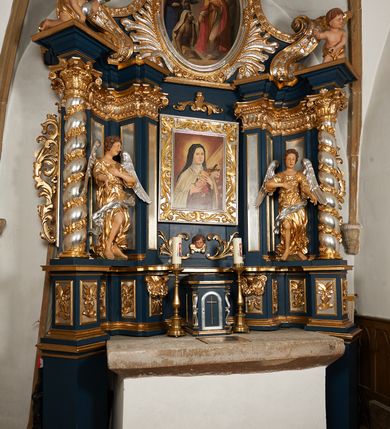Zdjęcie nr 13: Ołtarz architektoniczny, trójosiowy, jednokondygnacyjny ze zwieńczeniem, na cokole. Mensa kamienna z portatylem. Antepedium proste, bez dekoracji, kamienne. Tabernakulum prostopadłościenne, ujęte pilastrami ustawionymi pod kątem, ze zwróconymi przeciwstawnie wolutami, zamiast trzonów, zwieńczone niepełnym belkowaniem. Drzwiczki w formie stojącego prostokąta zamkniętego łukiem półkolistym z krzyżem w polu. Retabulum ujęte parami pilastrów i kolumn o skręconych trzonach oraz kapitelach kompozytowych, dźwigających niepełne belkowanie z wydatnym gzymsem, wyłamanym w osiach podpór, przerwane w polu środkowym. Na gzymsie para puttów trzymających arma Christi. Osie boczne ustawione pod kątem względem lica ołtarza, w nich ustawiony dwie figury stojących aniołów. W polu środkowym ołtarza rama w formie stojącego prostokąta mieszcząca obraz św. Teresy z Lisieux. Zwieńczenie ołtarza w kształcie owalnego medalionu ujętego wydatnymi spływami wolutowymi. W polu owalny obraz przedstawiający wskrzeszenie Piotrowina. Kondygnacja pierwsza nastawy ujęta uszami utworzonymi z akantu. Cokoły podpór ozdobione z trzech stron płycinami z dekoracją roślinną w postaci liści i kwiatów. Na osi pilastrów cokoły podparte potrójną wolutą i parą girland owocowych. Trzony pilastrów i osie boczne dekorowane płycinami. Trzony kolumn zdobione ornamentem roślinnym. W polu środkowym ołtarza rama obrazu dekorowana akantem; u jej spodu akant i uskrzydlona główka anielska, zwieńczona akantem z palmetą. W zwieńczeniu profilowana rama dekorowana miejscami akantem, ujęta muszlą; woluty i lizeny zdobione kampanulami. Struktura ołtarza polichromowana na ciemnogranatowo ze złoconymi i srebrzonymi elementami architektonicznymi i ornamentyką.