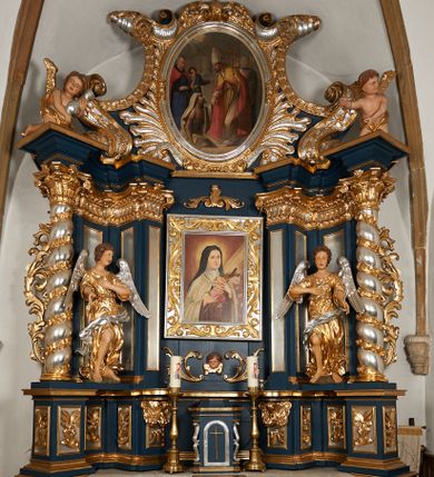 Zdjęcie nr 1: Ołtarz architektoniczny, trójosiowy, jednokondygnacyjny ze zwieńczeniem, na cokole. Mensa kamienna z portatylem. Antepedium proste, bez dekoracji, kamienne. Tabernakulum prostopadłościenne, ujęte pilastrami ustawionymi pod kątem, ze zwróconymi przeciwstawnie wolutami, zamiast trzonów, zwieńczone niepełnym belkowaniem. Drzwiczki w formie stojącego prostokąta zamkniętego łukiem półkolistym z krzyżem w polu. Retabulum ujęte parami pilastrów i kolumn o skręconych trzonach oraz kapitelach kompozytowych, dźwigających niepełne belkowanie z wydatnym gzymsem, wyłamanym w osiach podpór, przerwane w polu środkowym. Na gzymsie para puttów trzymających arma Christi. Osie boczne ustawione pod kątem względem lica ołtarza, w nich ustawiony dwie figury stojących aniołów. W polu środkowym ołtarza rama w formie stojącego prostokąta mieszcząca obraz św. Teresy z Lisieux. Zwieńczenie ołtarza w kształcie owalnego medalionu ujętego wydatnymi spływami wolutowymi. W polu owalny obraz przedstawiający wskrzeszenie Piotrowina. Kondygnacja pierwsza nastawy ujęta uszami utworzonymi z akantu. Cokoły podpór ozdobione z trzech stron płycinami z dekoracją roślinną w postaci liści i kwiatów. Na osi pilastrów cokoły podparte potrójną wolutą i parą girland owocowych. Trzony pilastrów i osie boczne dekorowane płycinami. Trzony kolumn zdobione ornamentem roślinnym. W polu środkowym ołtarza rama obrazu dekorowana akantem; u jej spodu akant i uskrzydlona główka anielska, zwieńczona akantem z palmetą. W zwieńczeniu profilowana rama dekorowana miejscami akantem, ujęta muszlą; woluty i lizeny zdobione kampanulami. Struktura ołtarza polichromowana na ciemnogranatowo ze złoconymi i srebrzonymi elementami architektonicznymi i ornamentyką.