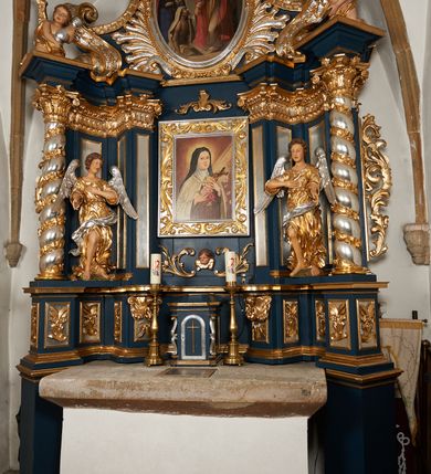 Zdjęcie nr 2: Ołtarz architektoniczny, trójosiowy, jednokondygnacyjny ze zwieńczeniem, na cokole. Mensa kamienna z portatylem. Antepedium proste, bez dekoracji, kamienne. Tabernakulum prostopadłościenne, ujęte pilastrami ustawionymi pod kątem, ze zwróconymi przeciwstawnie wolutami, zamiast trzonów, zwieńczone niepełnym belkowaniem. Drzwiczki w formie stojącego prostokąta zamkniętego łukiem półkolistym z krzyżem w polu. Retabulum ujęte parami pilastrów i kolumn o skręconych trzonach oraz kapitelach kompozytowych, dźwigających niepełne belkowanie z wydatnym gzymsem, wyłamanym w osiach podpór, przerwane w polu środkowym. Na gzymsie para puttów trzymających arma Christi. Osie boczne ustawione pod kątem względem lica ołtarza, w nich ustawiony dwie figury stojących aniołów. W polu środkowym ołtarza rama w formie stojącego prostokąta mieszcząca obraz św. Teresy z Lisieux. Zwieńczenie ołtarza w kształcie owalnego medalionu ujętego wydatnymi spływami wolutowymi. W polu owalny obraz przedstawiający wskrzeszenie Piotrowina. Kondygnacja pierwsza nastawy ujęta uszami utworzonymi z akantu. Cokoły podpór ozdobione z trzech stron płycinami z dekoracją roślinną w postaci liści i kwiatów. Na osi pilastrów cokoły podparte potrójną wolutą i parą girland owocowych. Trzony pilastrów i osie boczne dekorowane płycinami. Trzony kolumn zdobione ornamentem roślinnym. W polu środkowym ołtarza rama obrazu dekorowana akantem; u jej spodu akant i uskrzydlona główka anielska, zwieńczona akantem z palmetą. W zwieńczeniu profilowana rama dekorowana miejscami akantem, ujęta muszlą; woluty i lizeny zdobione kampanulami. Struktura ołtarza polichromowana na ciemnogranatowo ze złoconymi i srebrzonymi elementami architektonicznymi i ornamentyką.