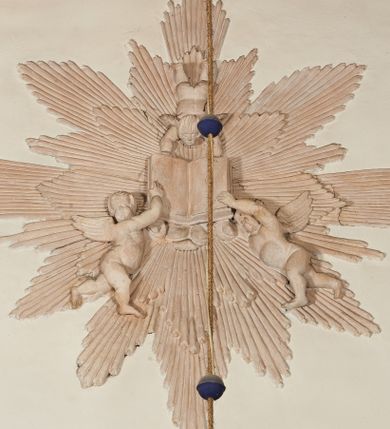 Zdjęcie nr 1: Naroża portalu do kaplicy ujęte wieńcami lauru i liśćmi akantu. Nad wejściem trzy putta trzymające otwartą księgę na tle monumentalnej glorii promienistej i obłoków. 