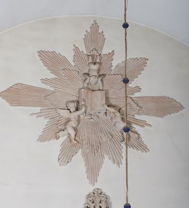 Zdjęcie nr 2: Naroża portalu do kaplicy ujęte wieńcami lauru i liśćmi akantu. Nad wejściem trzy putta trzymające otwartą księgę na tle monumentalnej glorii promienistej i obłoków. 