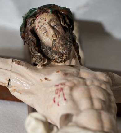 Zdjęcie nr 3: Krucyfiks drewniany, figura pełnoplastyczna, w pełni polichromowana. Ciało Chrystusa muskularne z silnie zaznaczoną anatomią klatki piersiowej i widocznymi śladami męki. Ramiona tworzące zarys litery „V”, prawa dłoń złożona w gest błogosławieństwa, lewa z wyprostowanym palcem wskazującym; stopy przebite jednym gwoździem. Głowa zwrócona w trzech czwartych w prawo, pochylona lekko do dołu. Twarz szczupła, pociągła, oczy zamknięte. Na głowie cierniowa korona, spod której ciekną krople krwi. Włosy i zarost ciemnobrązowe, kręcone. Perizonium białe, związane w węzeł na prawym boku postaci. U szczytu pionowej belki krzyża banderola z inskrypcją: „I.N./R.I.”