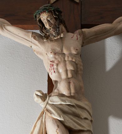 Zdjęcie nr 1: Krucyfiks drewniany, figura pełnoplastyczna, w pełni polichromowana. Ciało Chrystusa muskularne z silnie zaznaczoną anatomią klatki piersiowej i widocznymi śladami męki. Ramiona tworzące zarys litery „V”, prawa dłoń złożona w gest błogosławieństwa, lewa z wyprostowanym palcem wskazującym; stopy przebite jednym gwoździem. Głowa zwrócona w trzech czwartych w prawo, pochylona lekko do dołu. Twarz szczupła, pociągła, oczy zamknięte. Na głowie cierniowa korona, spod której ciekną krople krwi. Włosy i zarost ciemnobrązowe, kręcone. Perizonium białe, związane w węzeł na prawym boku postaci. U szczytu pionowej belki krzyża banderola z inskrypcją: „I.N./R.I.”