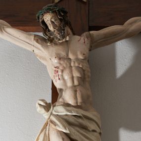 Zdjęcie nr 1: Krucyfiks drewniany, figura pełnoplastyczna, w pełni polichromowana. Ciało Chrystusa muskularne z silnie zaznaczoną anatomią klatki piersiowej i widocznymi śladami męki. Ramiona tworzące zarys litery „V”, prawa dłoń złożona w gest błogosławieństwa, lewa z wyprostowanym palcem wskazującym; stopy przebite jednym gwoździem. Głowa zwrócona w trzech czwartych w prawo, pochylona lekko do dołu. Twarz szczupła, pociągła, oczy zamknięte. Na głowie cierniowa korona, spod której ciekną krople krwi. Włosy i zarost ciemnobrązowe, kręcone. Perizonium białe, związane w węzeł na prawym boku postaci. U szczytu pionowej belki krzyża banderola z inskrypcją: „I.N./R.I.”