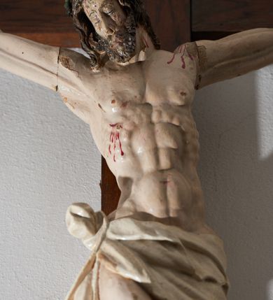 Zdjęcie nr 2: Krucyfiks drewniany, figura pełnoplastyczna, w pełni polichromowana. Ciało Chrystusa muskularne z silnie zaznaczoną anatomią klatki piersiowej i widocznymi śladami męki. Ramiona tworzące zarys litery „V”, prawa dłoń złożona w gest błogosławieństwa, lewa z wyprostowanym palcem wskazującym; stopy przebite jednym gwoździem. Głowa zwrócona w trzech czwartych w prawo, pochylona lekko do dołu. Twarz szczupła, pociągła, oczy zamknięte. Na głowie cierniowa korona, spod której ciekną krople krwi. Włosy i zarost ciemnobrązowe, kręcone. Perizonium białe, związane w węzeł na prawym boku postaci. U szczytu pionowej belki krzyża banderola z inskrypcją: „I.N./R.I.”