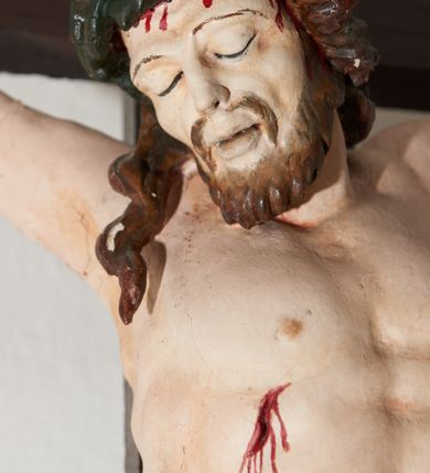 Zdjęcie nr 2: Krucyfiks drewniany, figura pełnoplastyczna, polichromowana. Ciało Chrystusa esowato wygięte o delikatnie zaznaczonej muskulaturze i widocznych śladach męki. Ramiona tworzą zarys litery „V”, stopy przebite jednym gwoździem. Głowa przechylona w prawo, złożona na prawym ramieniu postaci; oczy zamknięte, usta lekko rozwarte. Na głowie cierniowa korona, spod której ciekną krople krwi. Perizonium krótkie, srebrzone owinięte, wokół złoconego sznura i przeplecione pomiędzy udami postaci. U szczytu pionowej belki krzyża biały titulus z inskrypcją „I.N./R.I.” 