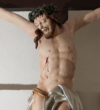 Zdjęcie nr 1: Krucyfiks drewniany, figura pełnoplastyczna, polichromowana. Ciało Chrystusa esowato wygięte o delikatnie zaznaczonej muskulaturze i widocznych śladach męki. Ramiona tworzą zarys litery „V”, stopy przebite jednym gwoździem. Głowa przechylona w prawo, złożona na prawym ramieniu postaci; oczy zamknięte, usta lekko rozwarte. Na głowie cierniowa korona, spod której ciekną krople krwi. Perizonium krótkie, srebrzone owinięte, wokół złoconego sznura i przeplecione pomiędzy udami postaci. U szczytu pionowej belki krzyża biały titulus z inskrypcją „I.N./R.I.” 
