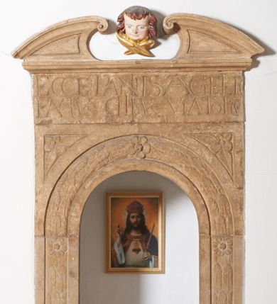 Zdjęcie nr 1: Sakramentarium w formie półkoliście zamkniętej niszy w kamiennym obramieniu o architektonicznych formach. Nisza ujęta arkadą  ustawioną na cokole dekorowanym rautami. Arkada wypełniona od frontu girlandą oraz trzema kwiatami. W przyłuczach trójkątne pola wypełnione kwiatami. Powyżej fryz z inskrypcją: „ECCE PANIS A(N)GELOR[UM] / FACTUS CIBUS VIATORU(M) / MM”. W zwieńczeniu wolutowo zawinięte odcinki przyczłóka. Pomiędzy nimi drewniana, polichromowana i złocona uskrzydlona główka anielska.